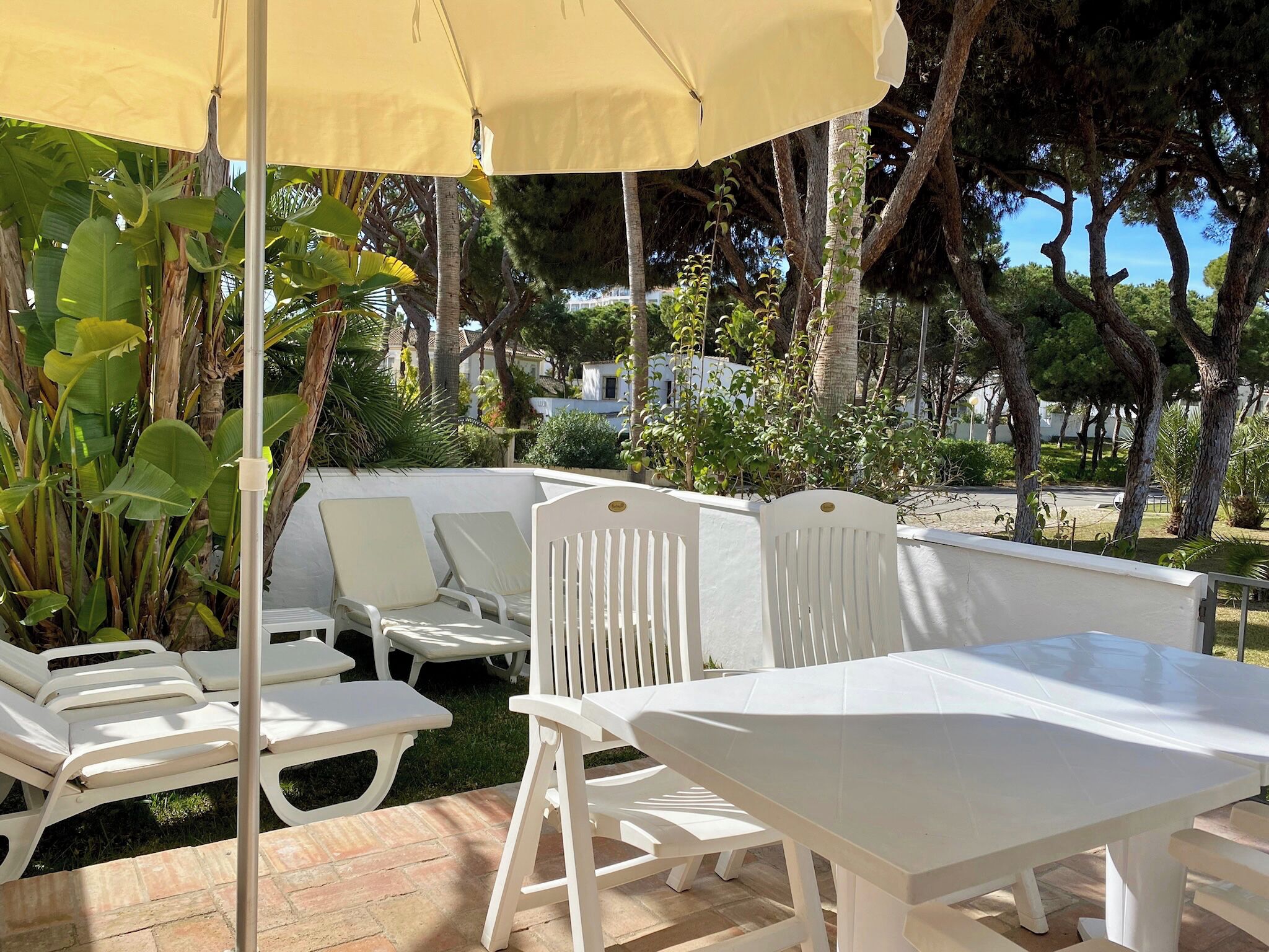 An outdoor patio with white lounge chairs and a table under a yellow umbrella, surrounded by lush greenery and trees.