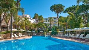 A swimming pool with lounge chairs and umbrellas, surrounded by palm trees and buildings. - Vale Do Garrão Villas (Loulé)