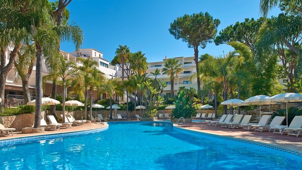 A swimming pool with lounge chairs and umbrellas, surrounded by palm trees and buildings. - Vale Do Garrão Villas (Loulé)