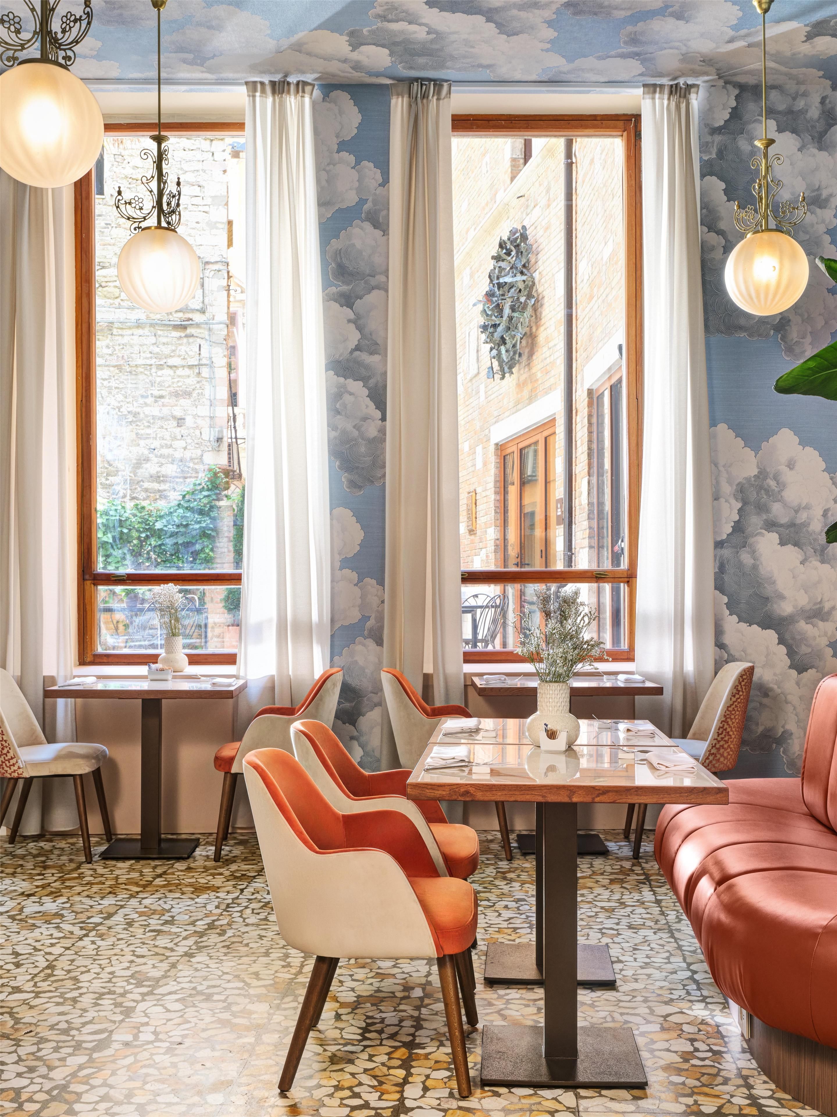 A dining area with a table set for four, surrounded by chairs with a mix of colors, large windows with curtains, and a ceiling with a cloud mural.