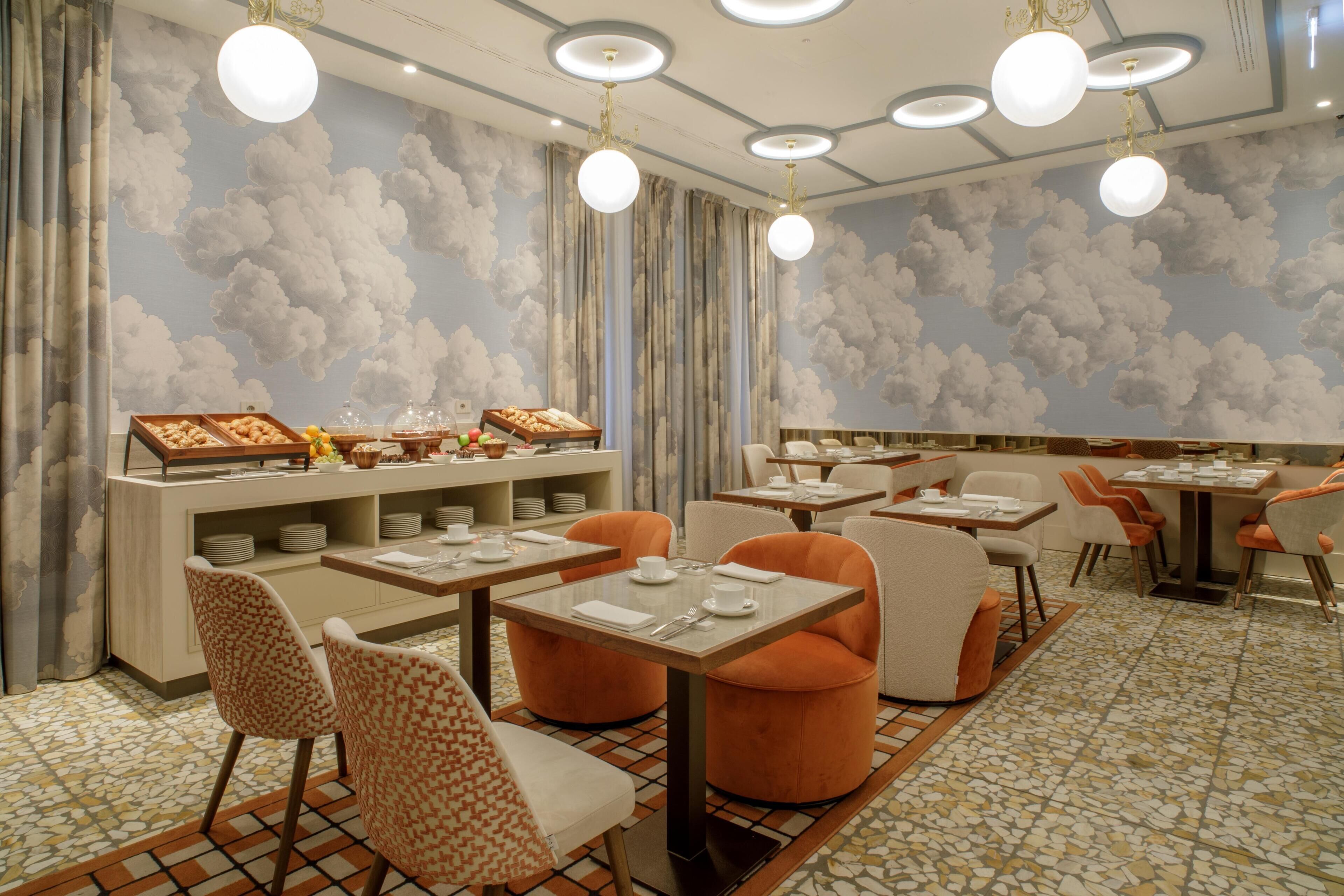 A restaurant with round tables, orange chairs, and a ceiling with cloud-themed lighting.