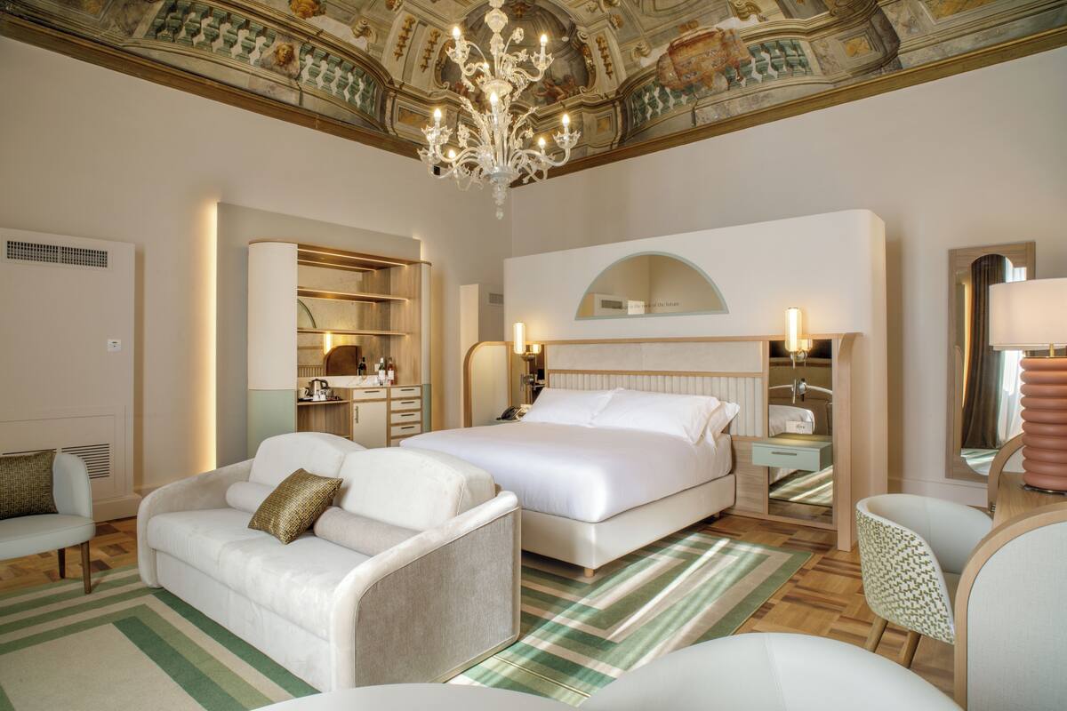 A hotel room with a large bed, a sitting area with a sofa and armchair, a vanity with a mirror, and a chandelier.