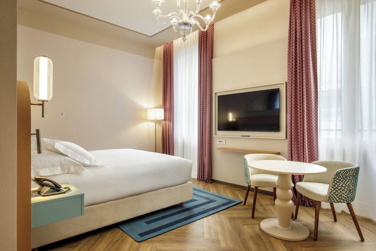 A hotel room with a bed, bedside table with a phone, a TV, a small round table with chairs, and a chandelier.