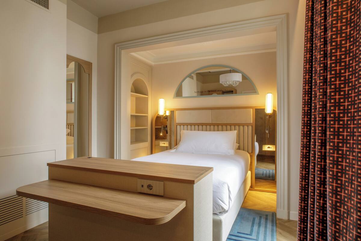 A hotel room with a bed, bedside tables, a mirror, and a view into the hallway.