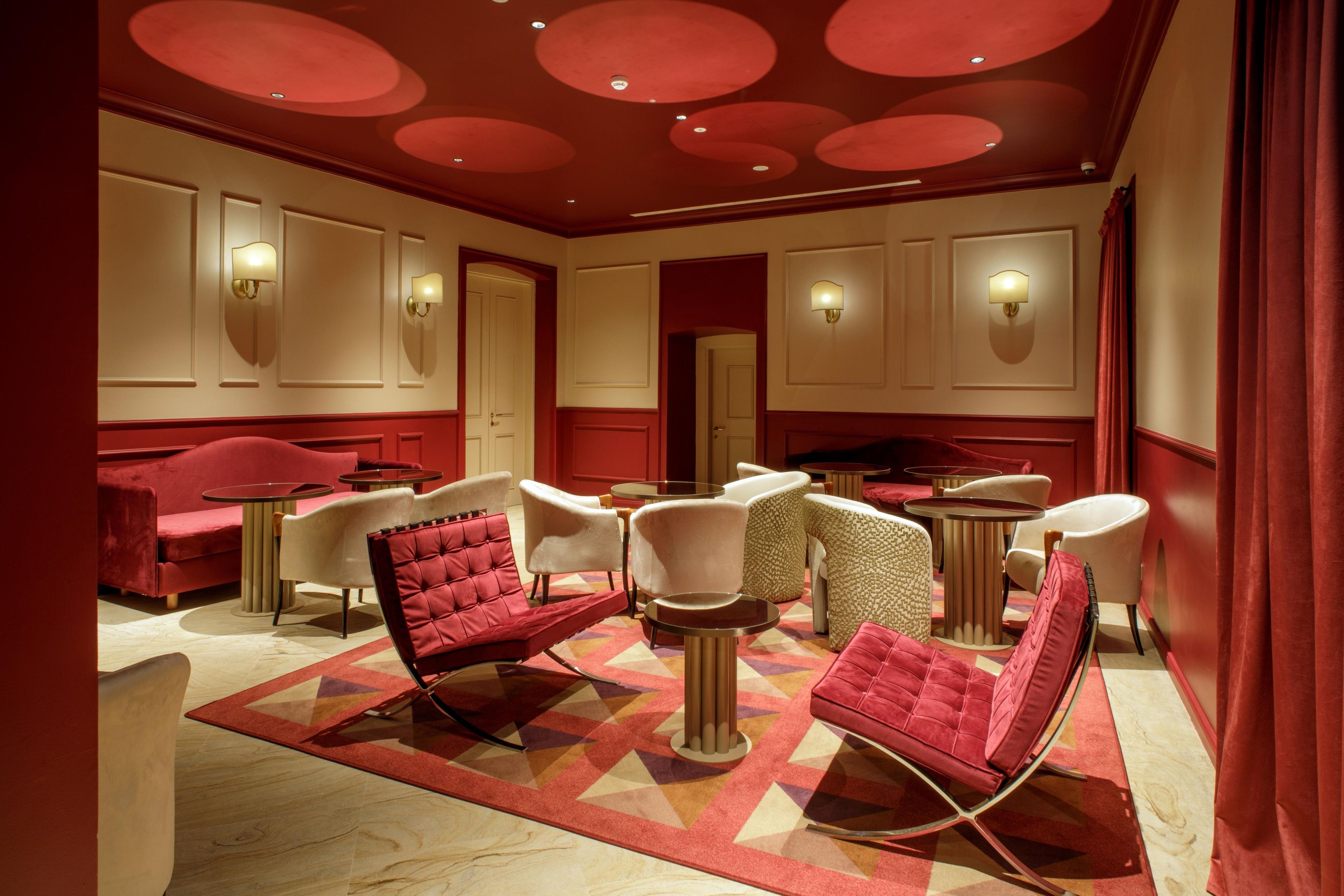 A hotel room with a red and beige color scheme, featuring a red sofa, white armchairs, and a patterned rug.