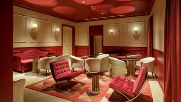A hotel room with a red and beige color scheme, featuring a red sofa, white armchairs, and a patterned rug.