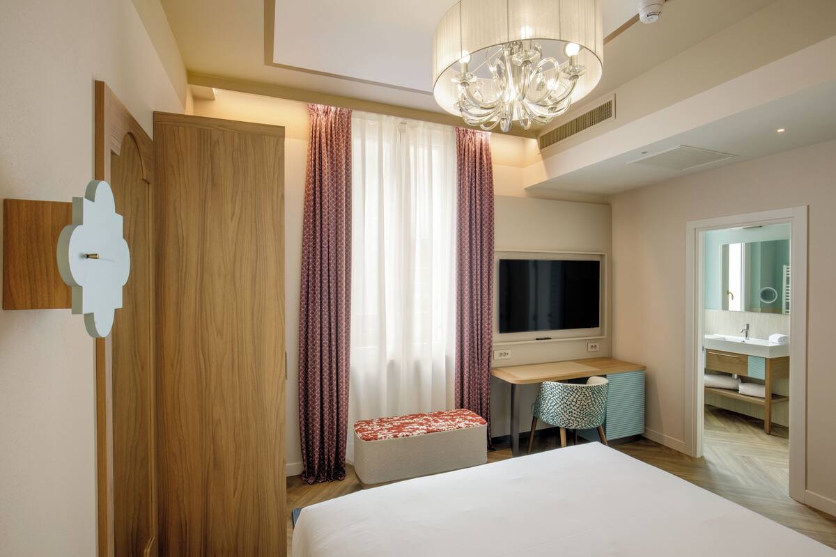 A hotel room with a bed, a desk, a TV, a chair, and a bathroom visible through an open door.
