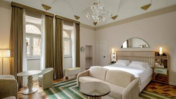 Habitación de hotel con una cama grande, una zona de estar con una silla y una mesita, un espejo sobre la cama y una araña.