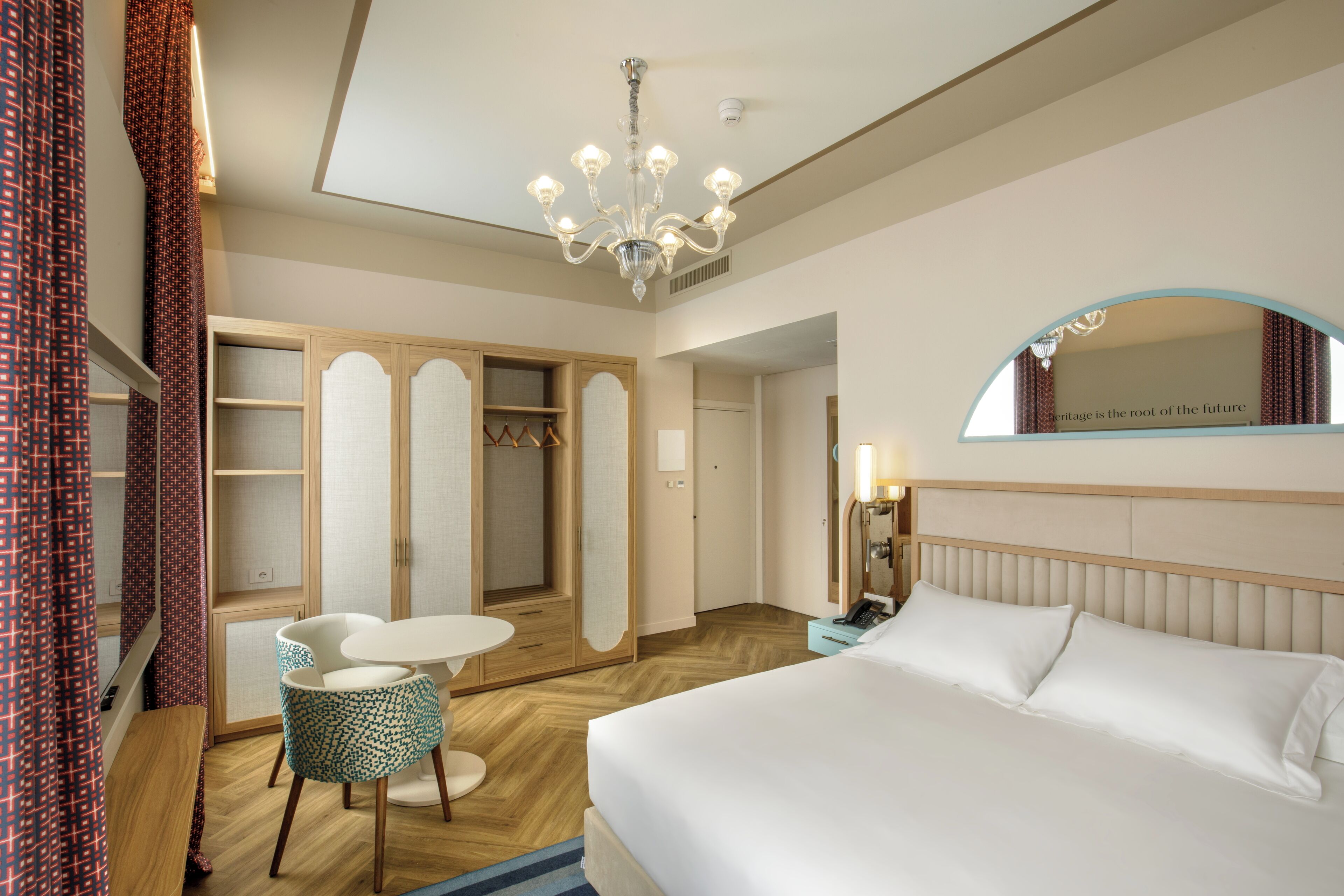 A hotel room with a bed, a desk with a chair, a wardrobe, a mirror, and a chandelier.