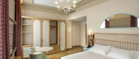A hotel room with a bed, a desk with a chair, a wardrobe, a mirror, and a chandelier.