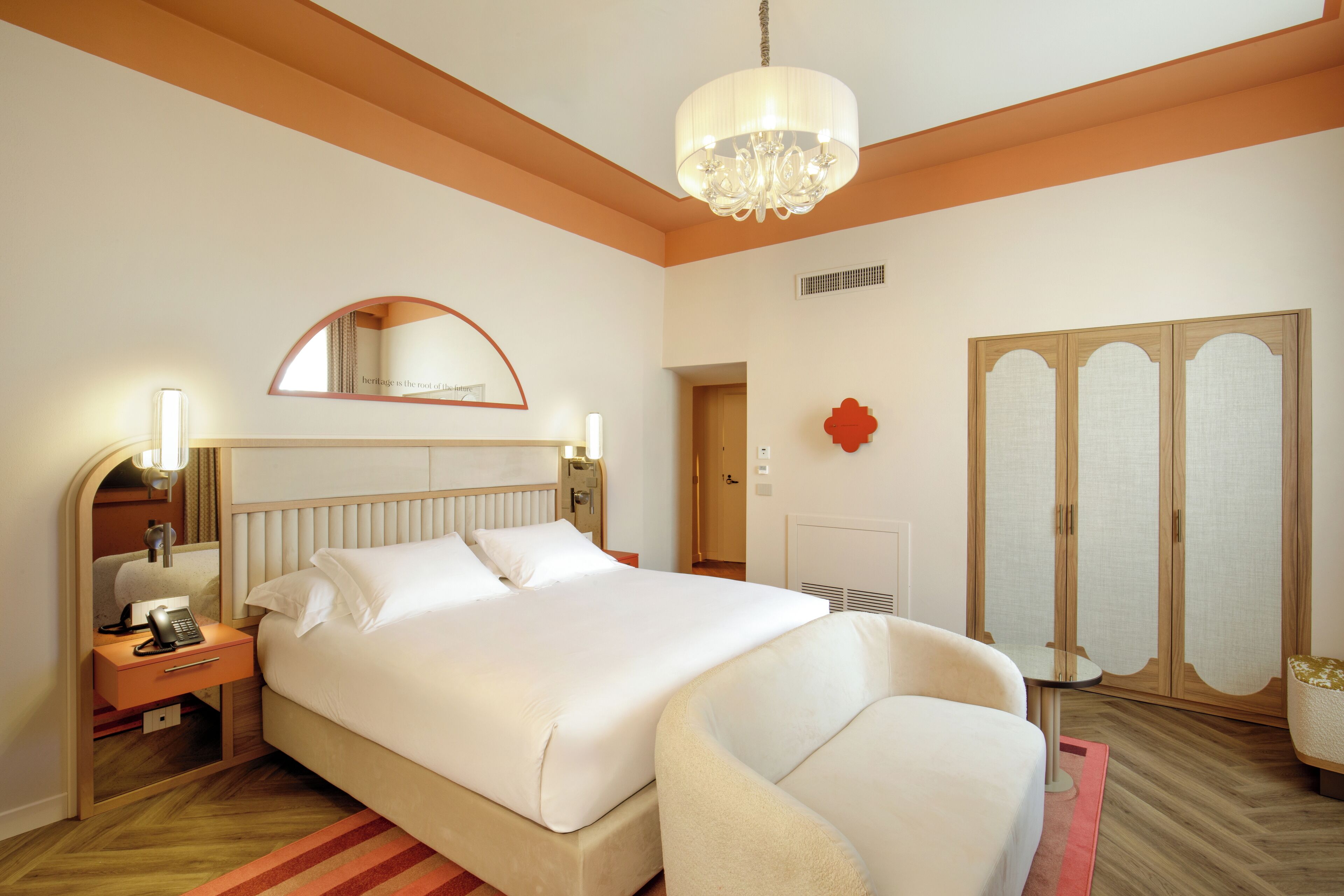 A hotel room with a bed, bedside table, lamp, mirror, wardrobe, and a chandelier.