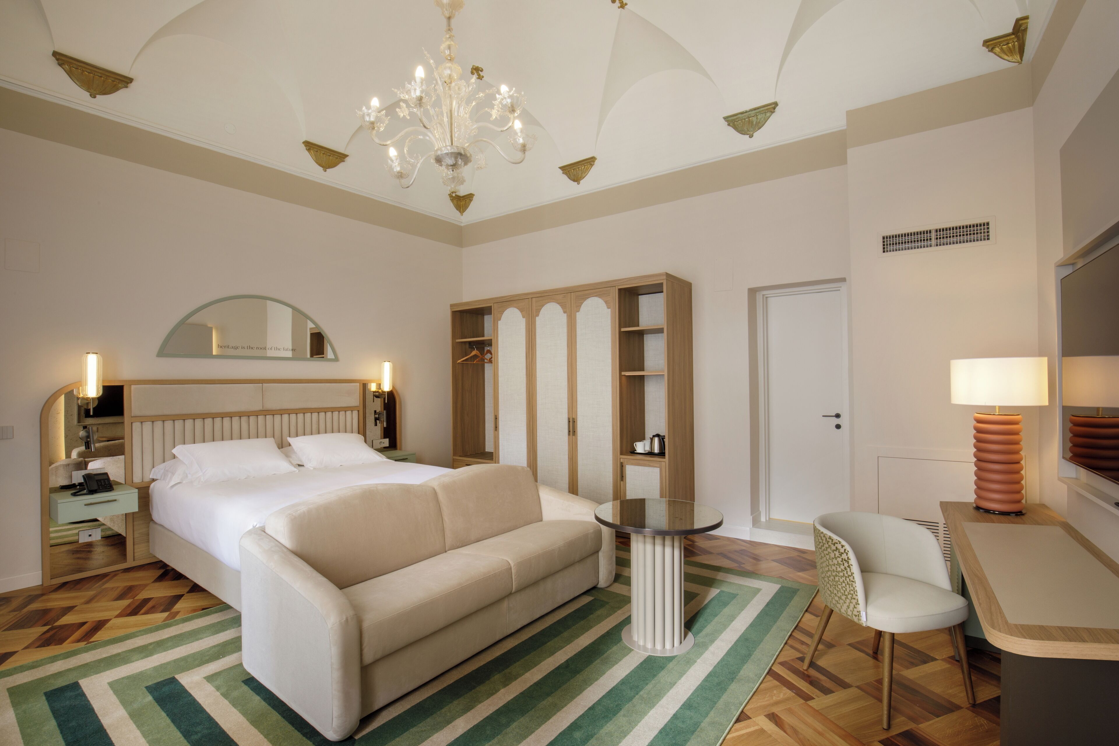 A hotel room with a bed, a sofa, a small table, a chair, a wardrobe, a mirror, and a chandelier.