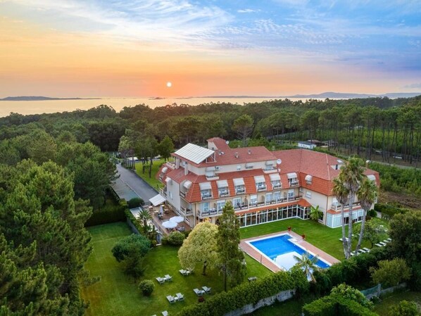 A large, multi-story building with a swimming pool, surrounded by lush greenery and a view of the sunset over a body of water.