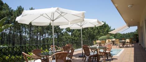 Un patio al aire libre con mobiliario de mimbre y sombrillas blancas, con vista a una piscina y zona boscosa.