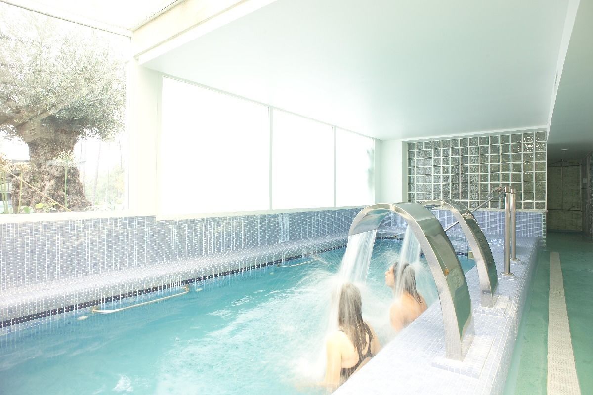 A swimming pool with a waterfall feature, surrounded by a tiled wall and large windows.