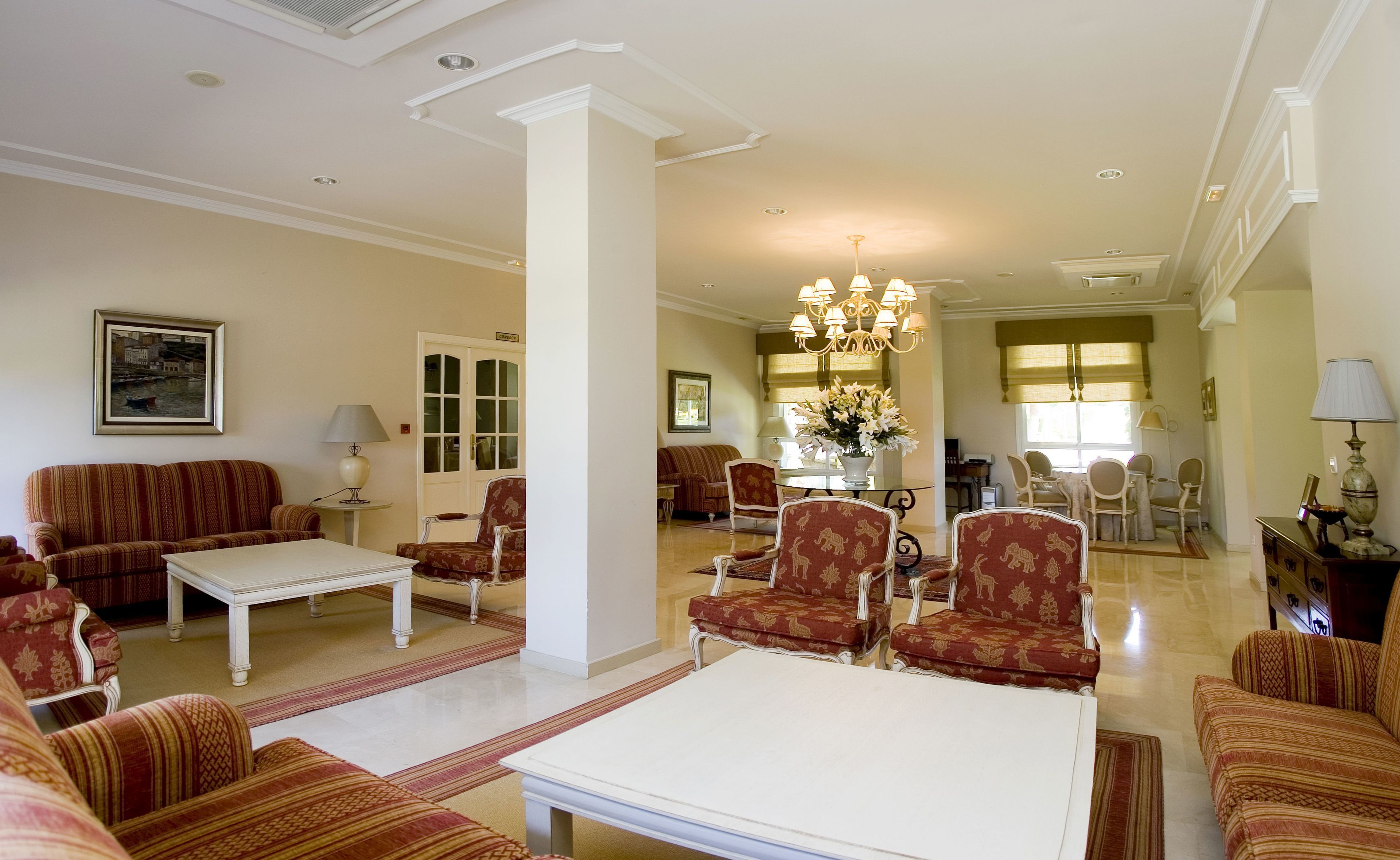 A spacious living room with a white coffee table, striped sofas, and patterned armchairs. A chandelier hangs from the ceiling, and a dining area is visible in the background.