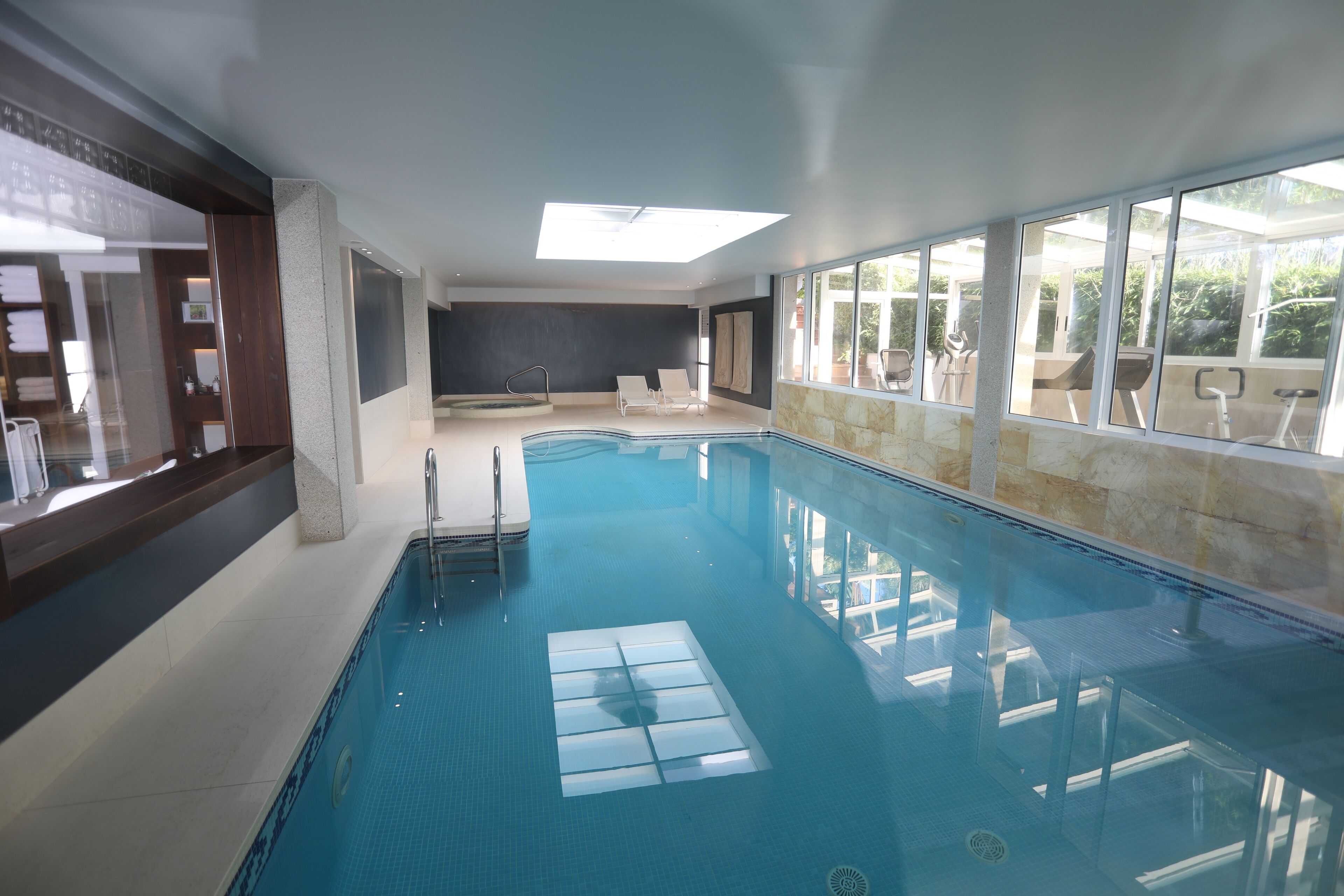 An indoor swimming pool with clear glass walls, a sunlit ceiling, and a view of outdoor exercise equipment.