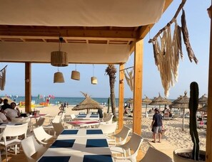 Un área de estar al aire libre con mesas y sillas, una pérgola de madera y vistas a la playa.