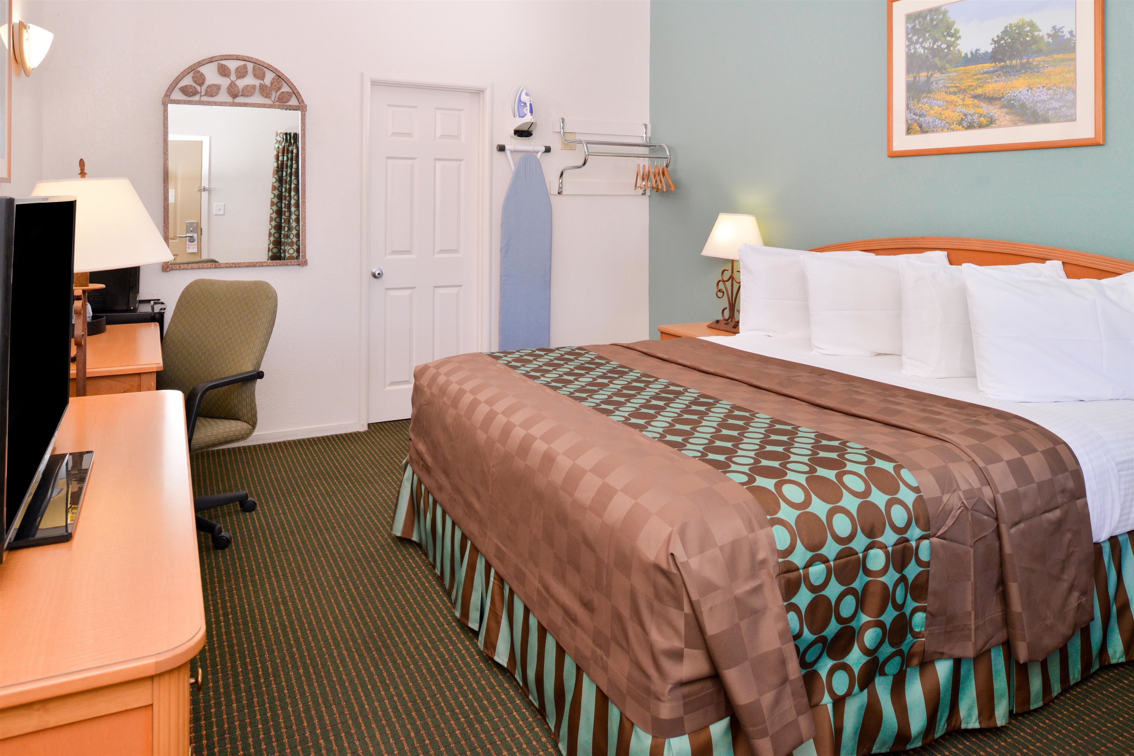 A hotel room with a bed, a desk with a lamp, a chair, a mirror, and a door.
