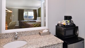 A hotel bathroom with a granite countertop sink, a mirror, a small refrigerator, and a coffee maker.