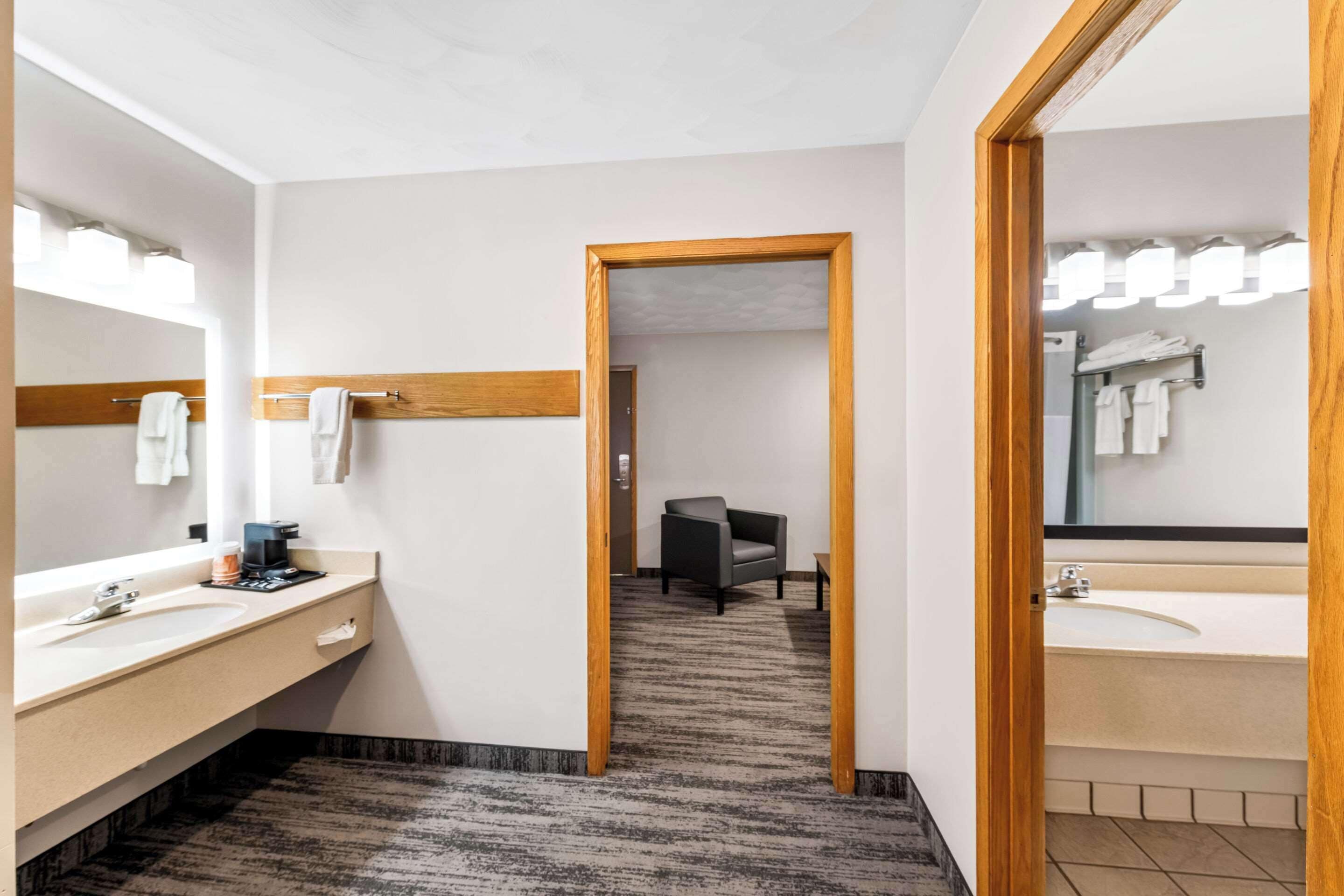 A modern bathroom with a large mirror, two sinks, a chair, and a wooden door frame.