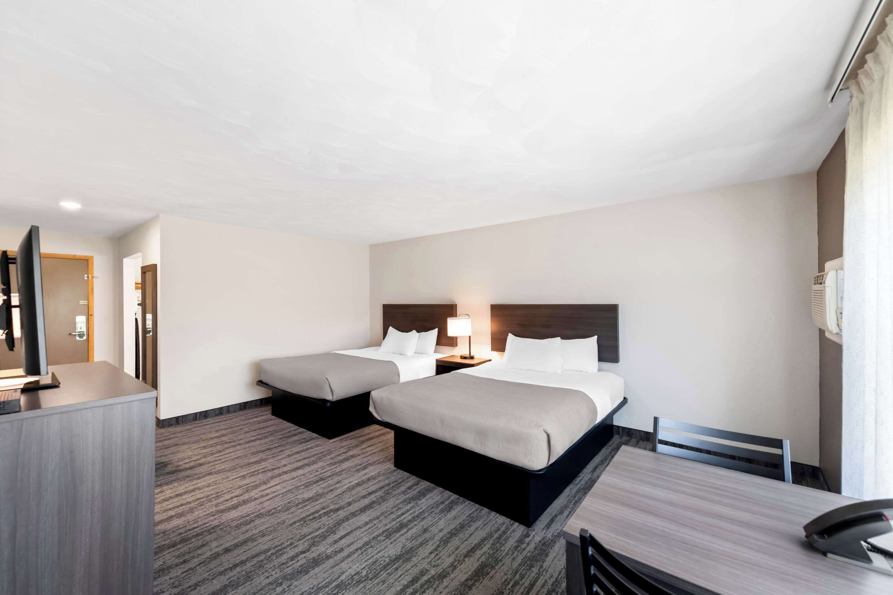 A hotel room with two beds, a desk, a chair, a TV, and a lamp.