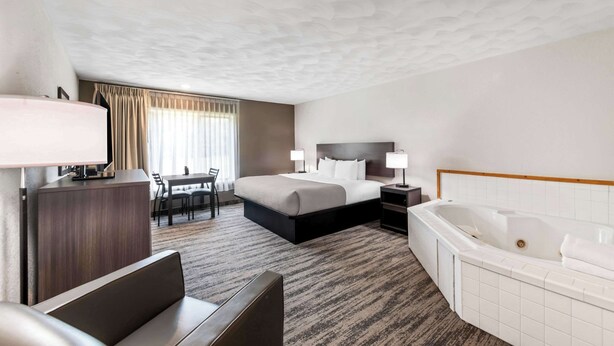 A hotel room with a bed, a desk, a chair, a desk lamp, a bedside table with a lamp, and a large bathtub. - Quality Inn & Suites Munising (Munising)