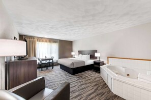 Een hotelkamer met een bed, een bureau, een stoel, een bureaulamp, een nachtkastje met lamp en een groot bad.