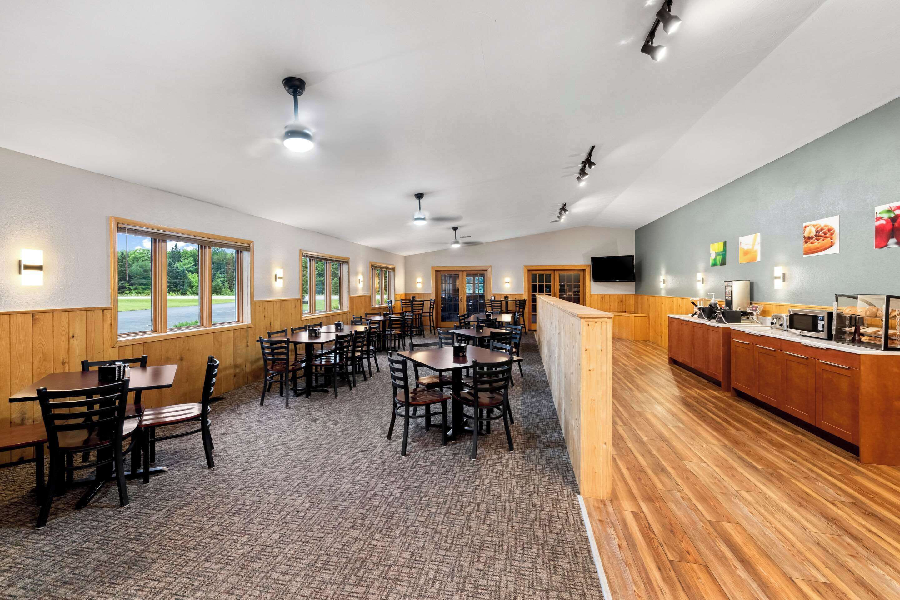 Ein Restaurant mit Holztischen und -stühlen, einer Küchenzeile und großen Fenstern mit Blick auf Grünflächen.