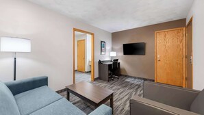 A hotel room with a sofa, a small table, a desk with a chair, a TV mounted on the wall, and a wooden door. - Quality Inn & Suites Munising (Munising)