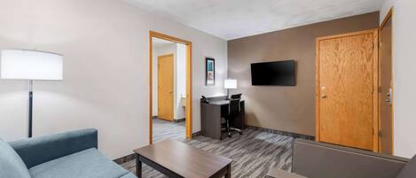 A hotel room with a sofa, a small table, a desk with a chair, a TV mounted on the wall, and a wooden door.