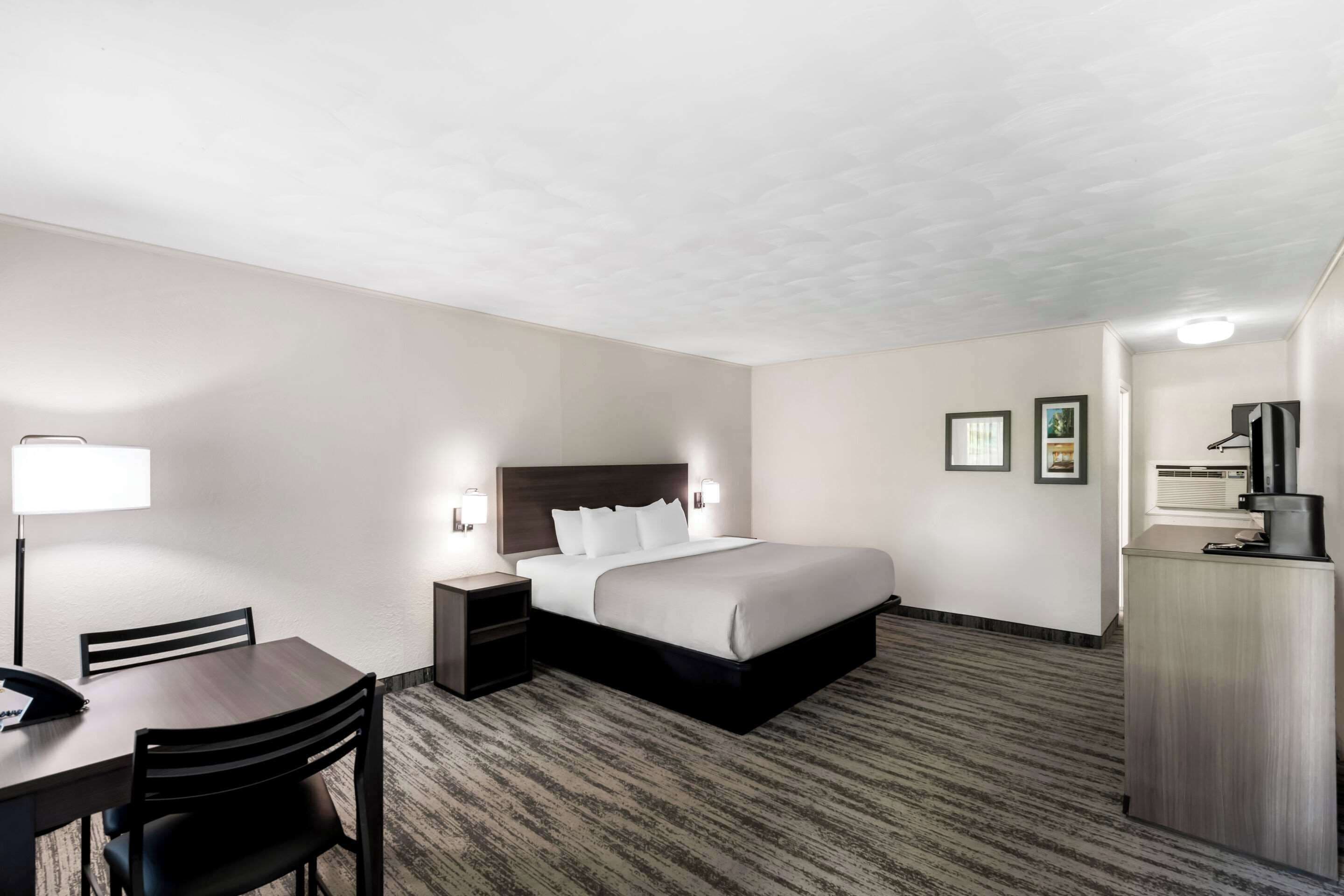 A hotel room with a bed, a desk with a chair, a nightstand, a dresser, and a lamp.