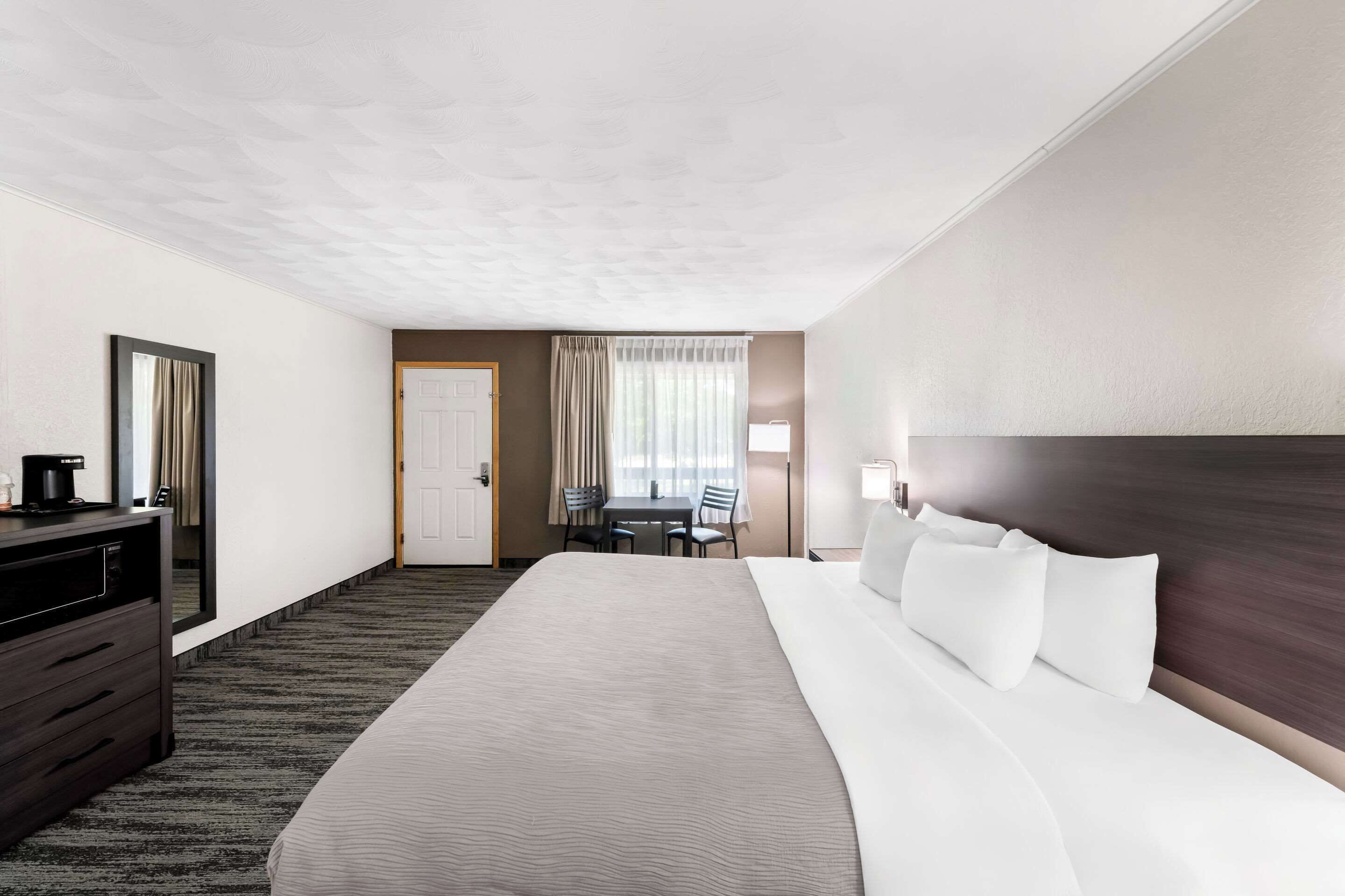 Quarto de hotel com uma cama grande, cômoda, escrivaninha com cadeiras e janela com cortinas.