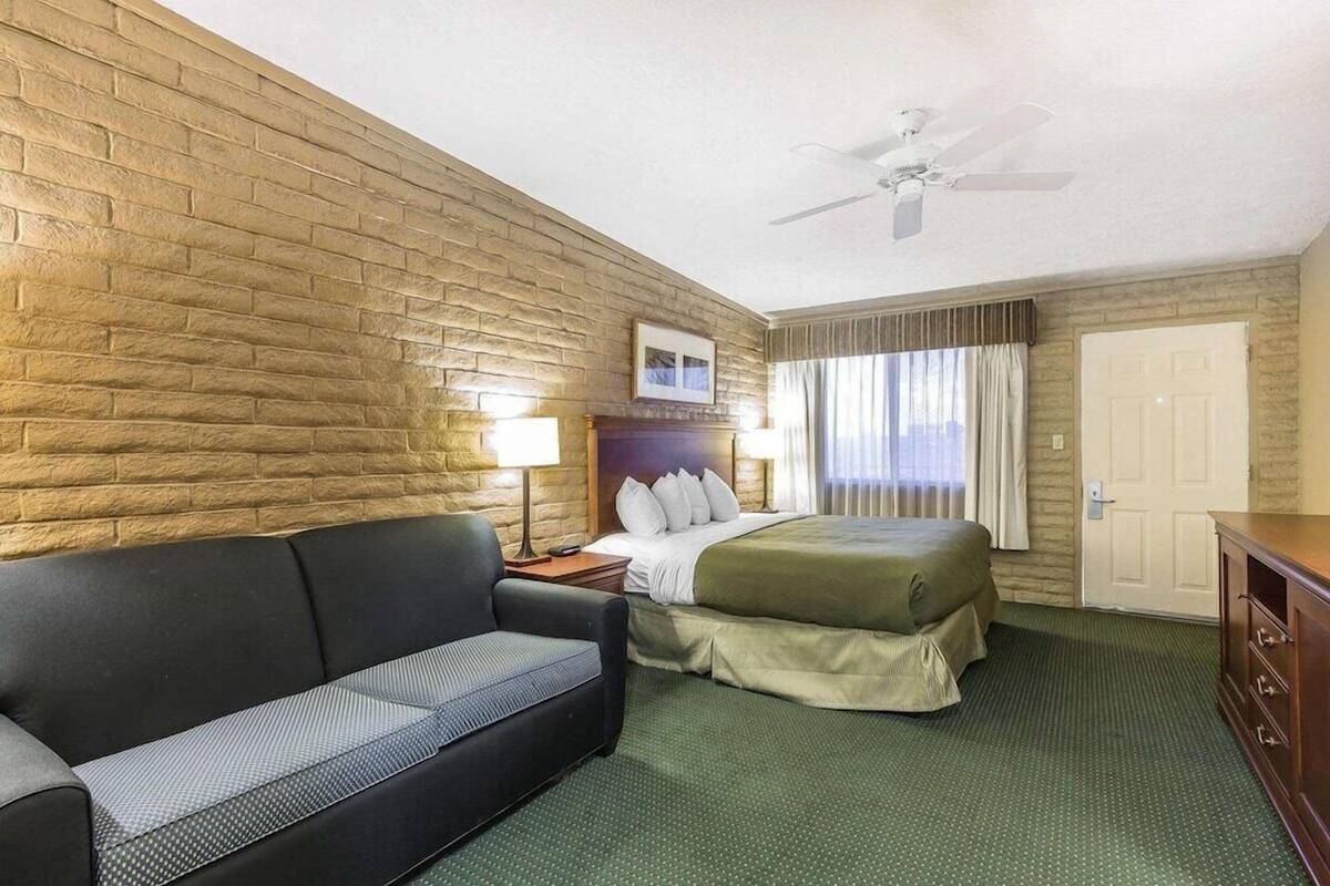 A hotel room with a brick wall, a bed, a sofa, a ceiling fan, and a door.