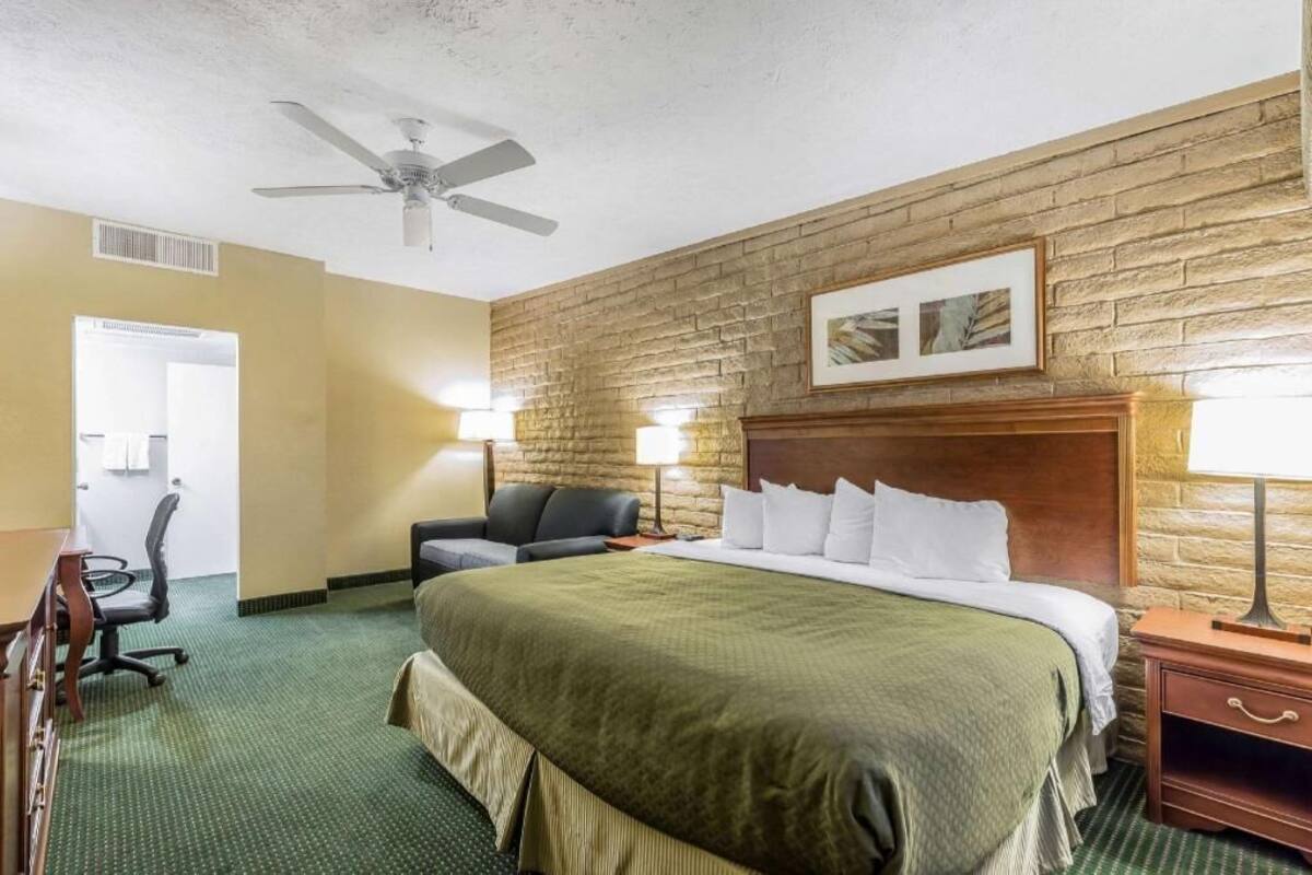 A hotel room with a large bed, a desk, a chair, a ceiling fan, and a brick wall.