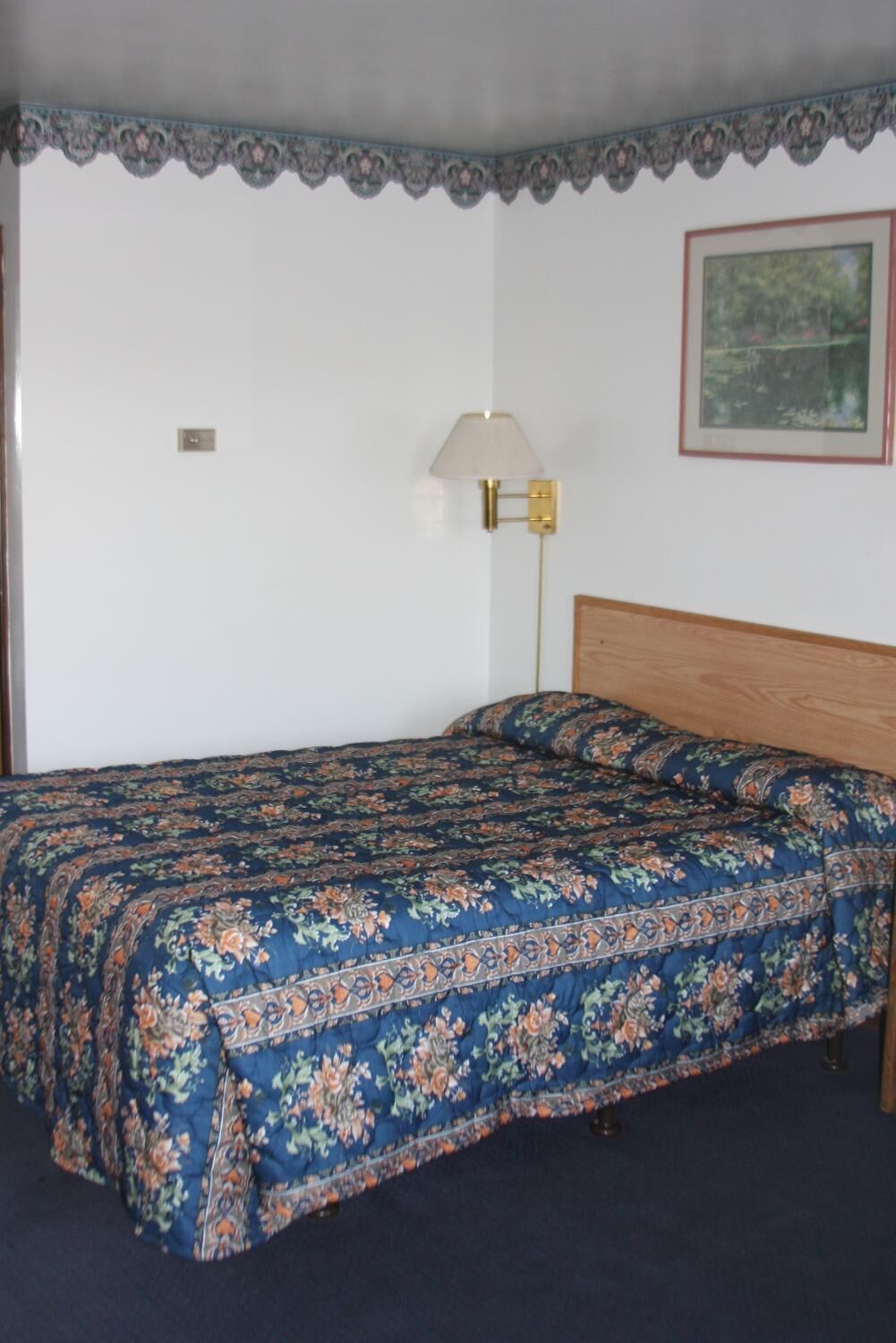Un dormitorio con cabecera de madera, edredón estampado de flores, lámpara de pared e imagen enmarcadas en la pared.