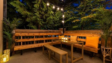 An outdoor seating area with wooden benches and tables, surrounded by greenery and string lights.