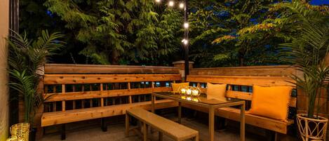 Un área de estar al aire libre con bancos y mesas de madera, rodeada de follaje e iluminada con luces colgantes.