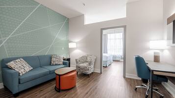 Una habitación de hotel moderna con un sofá azul, un reposapiés naranja pequeño, un sillón estampado, un escritorio con una silla y una cama con una colcha blanca.