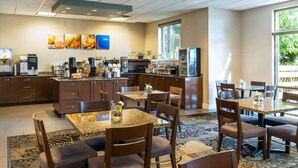 A hotel dining area with a kitchen in the background, a large window, and a carpeted floor.