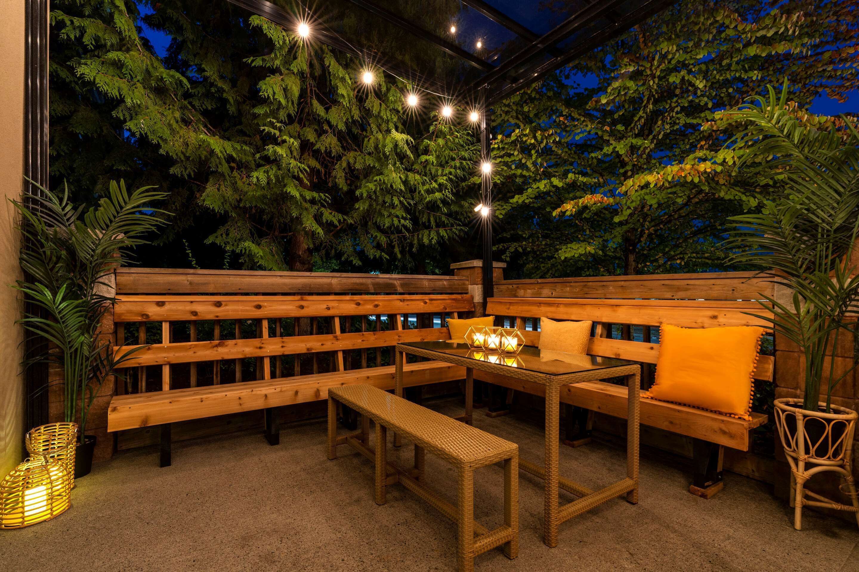 An outdoor seating area with wooden benches and tables, surrounded by greenery and string lights.