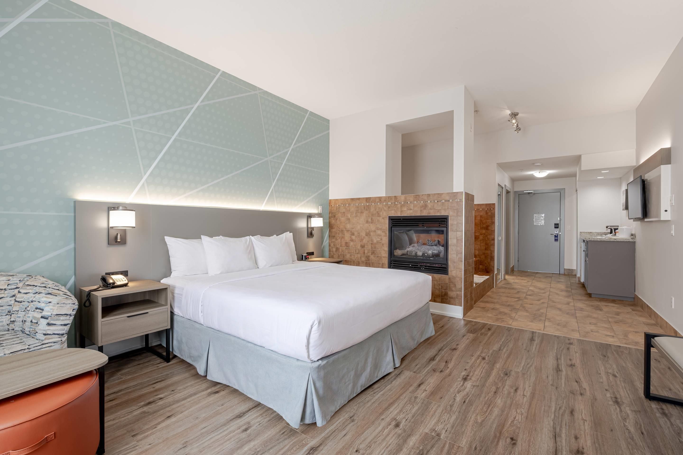 A hotel room with a large bed, bedside tables, a desk, a chair, a TV, a fireplace, and a bathroom visible through an open door.