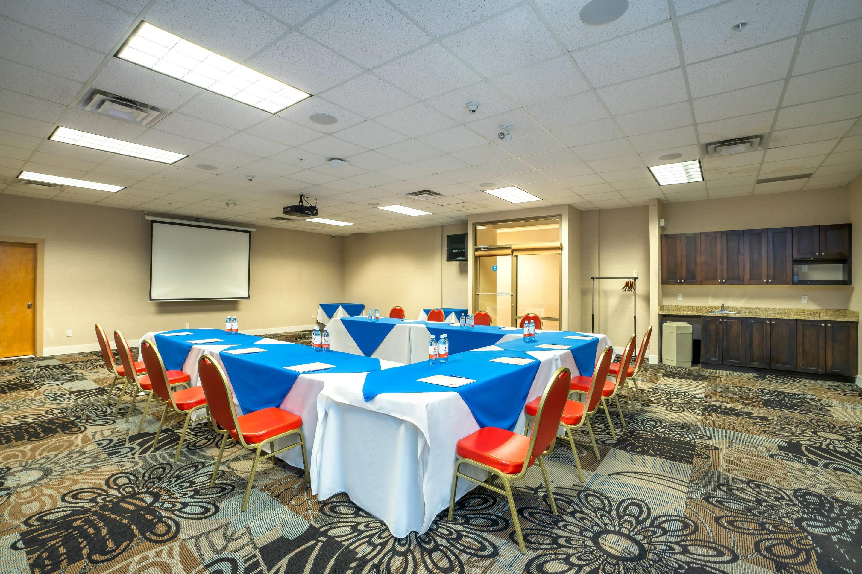 A conference room with tables set for meetings, equipped with chairs, a projector screen, and a kitchenette area.