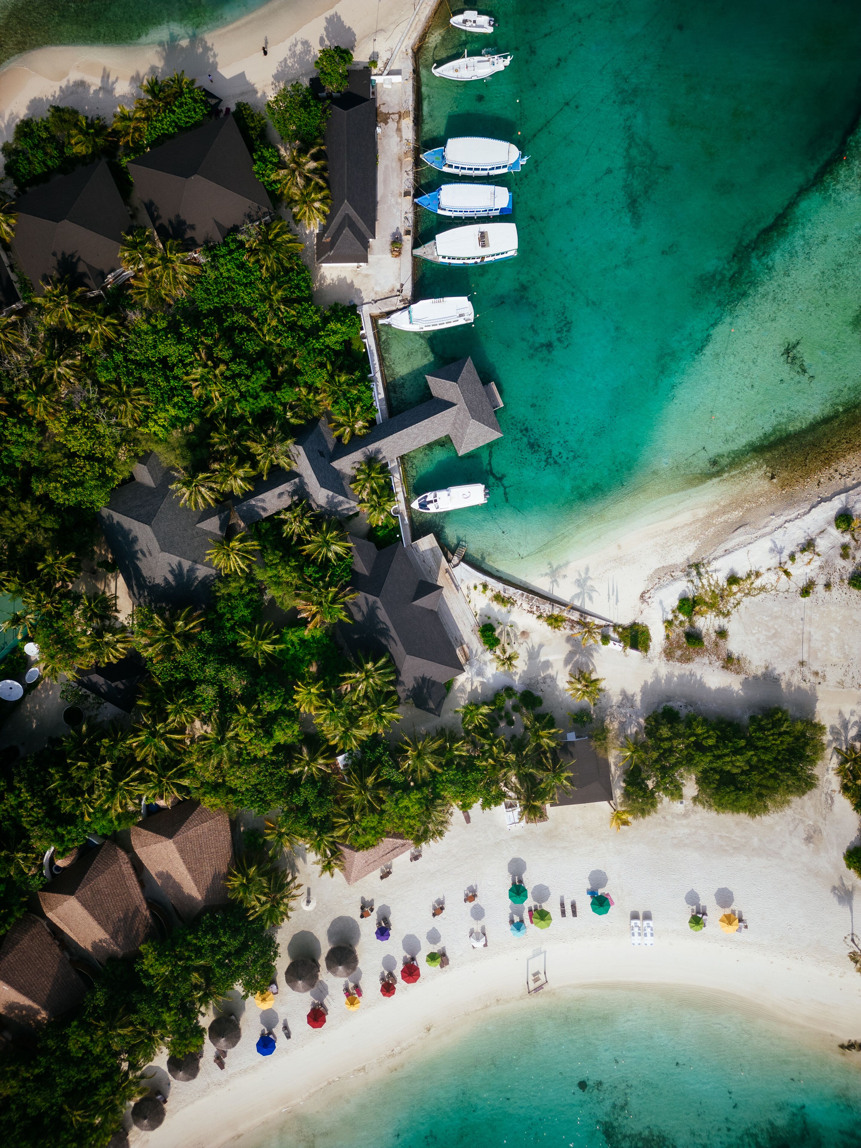 An aerial view of a resort with multiple boats docked at the pier, a beach with umbrellas, and lush greenery.