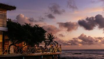 A coastal building with a balcony overlooking the sea, featuring a palm tree and a thatched roof.