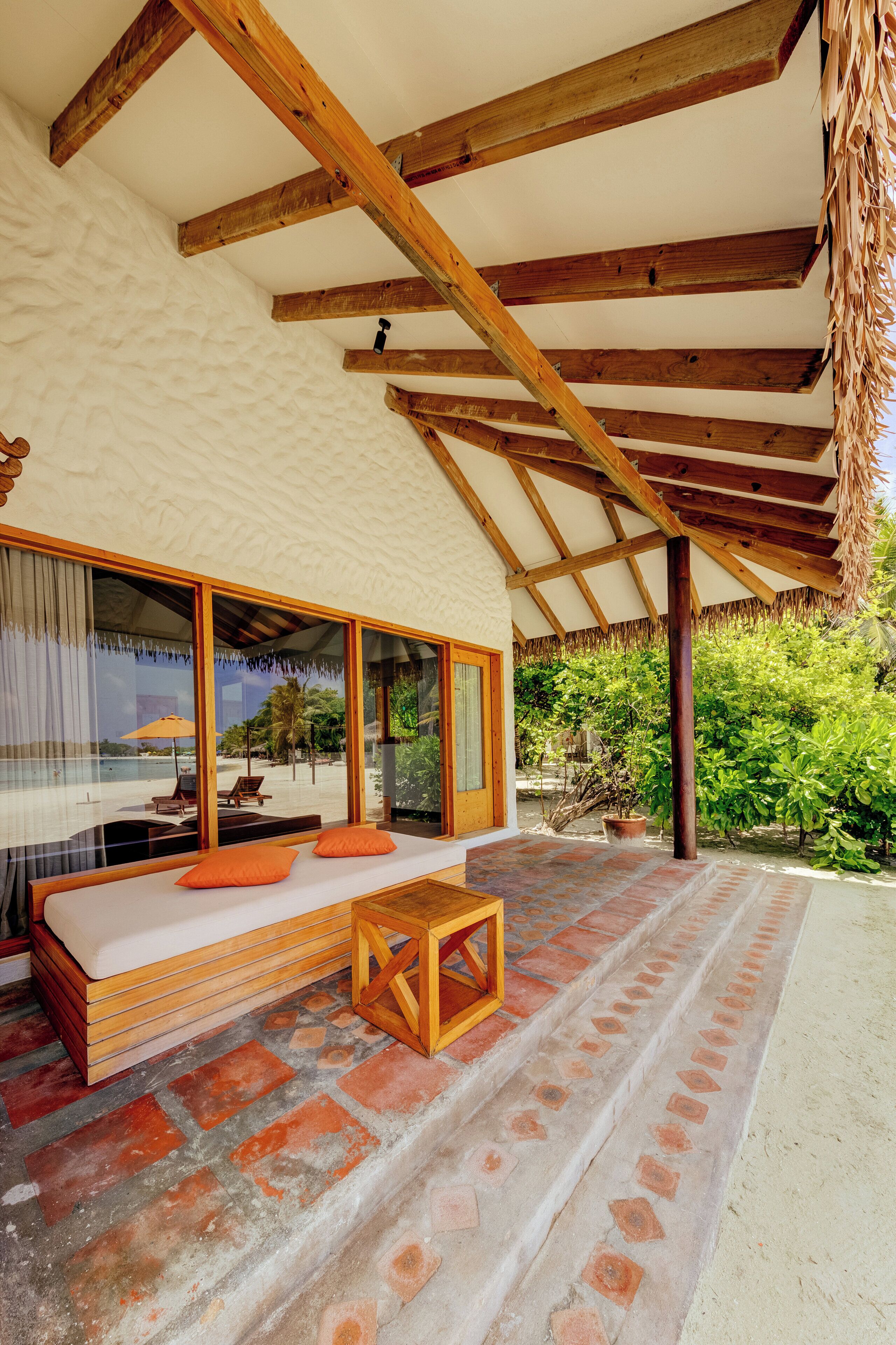 A covered outdoor seating area with a sofa and cushions, a wooden table, and a view of the beach through large windows.