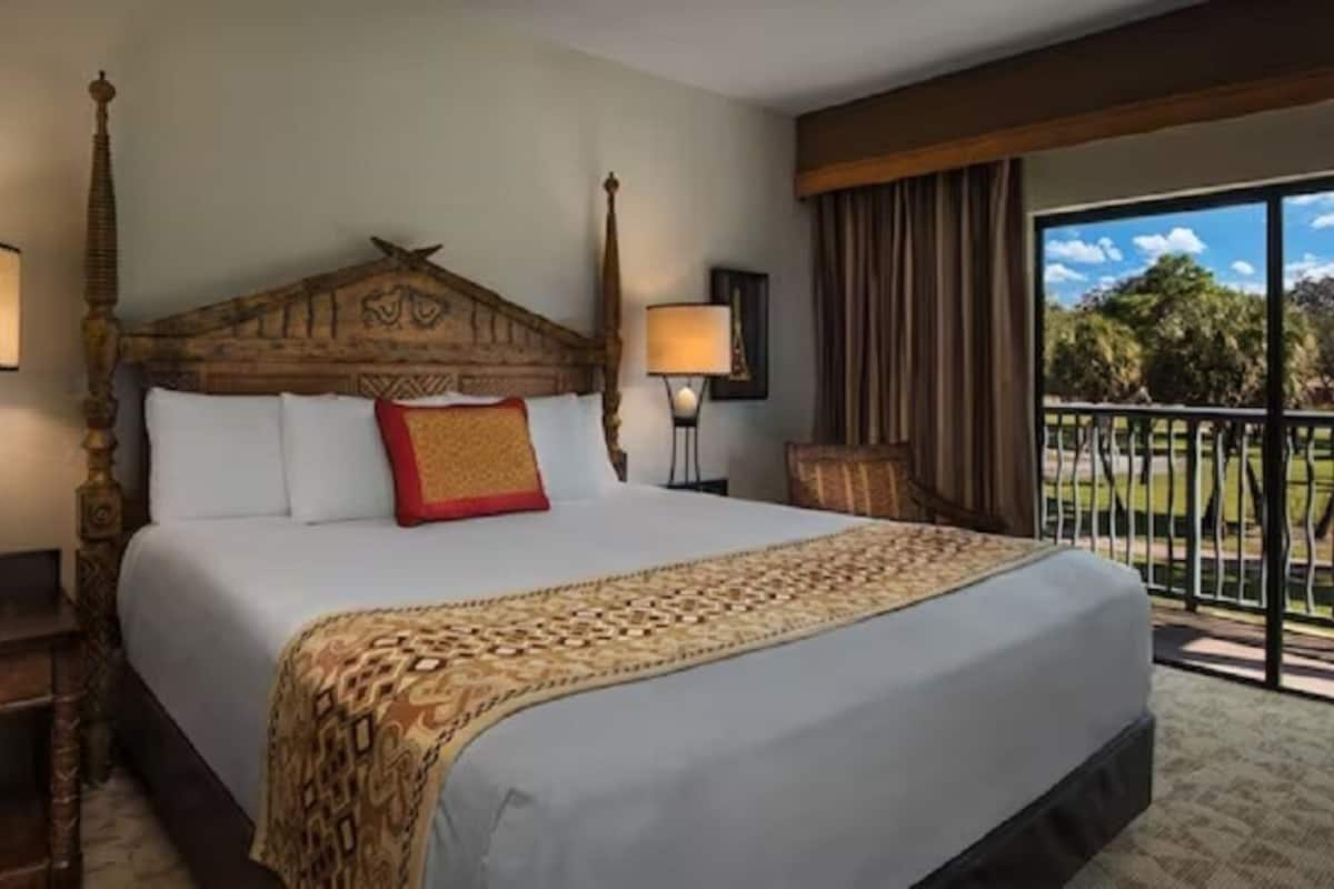 A hotel room with a large bed, two bedside tables, a window with a view of greenery, and a balcony.