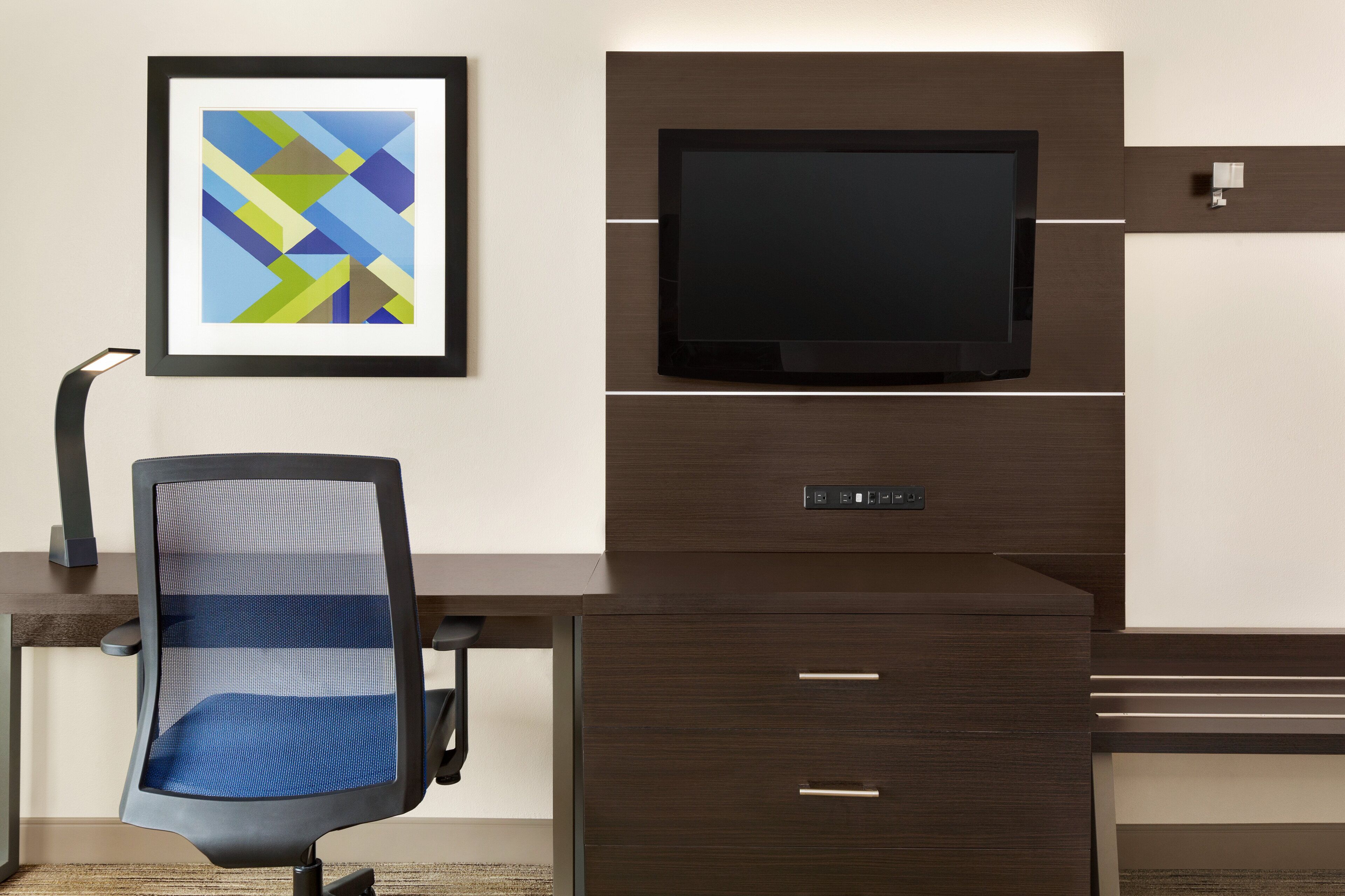 A modern office setup with a desk, chair, mounted TV, and a framed abstract artwork.