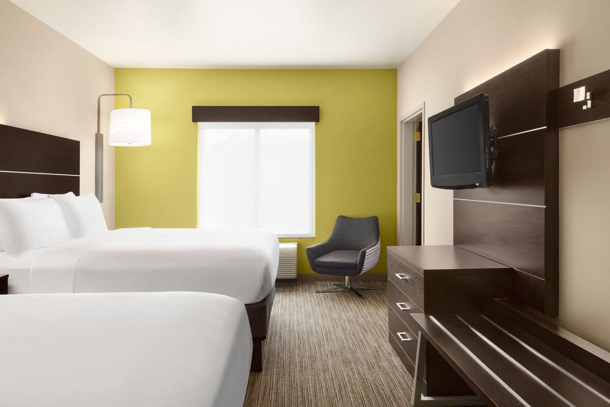 A hotel room with two beds, a chair, a TV, and a window with blinds.