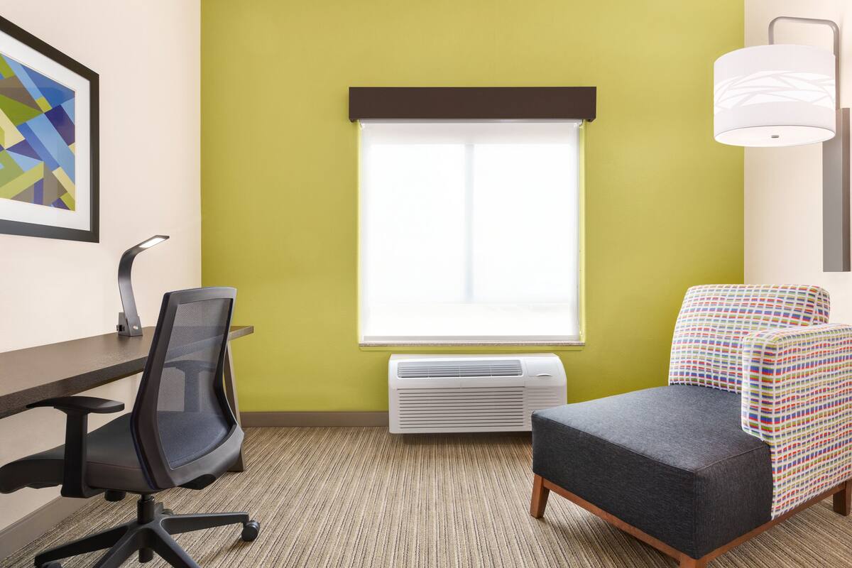 Una habitación con escritorio, silla de oficina, aire acondicionado, ventana con persianas y una silla de colores.